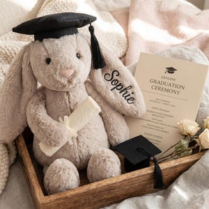 May include: A soft, light brown bunny toy wearing a black graduation cap and tassel, holding a scroll. A graduation announcement and miniature cap are in a wooden tray. The bunny's ear is embroidered with the name "Sophie".