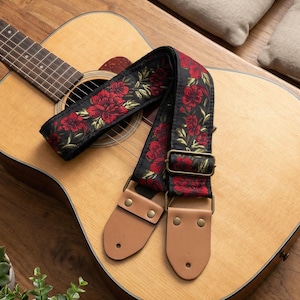 May include: Acoustic guitar with a black guitar strap featuring a red floral pattern. The strap has brown leather ends and metal hardware. The guitar is light brown and sits on a wooden surface.