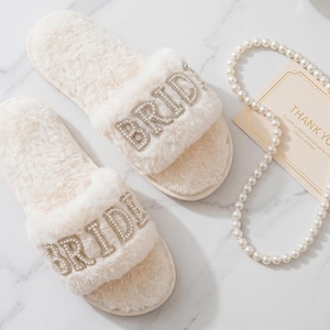 May include: Ivory-colored, fluffy slide slippers with the word "BRIDE" spelled out in sparkling rhinestones. A pearl necklace and a "Thank You" card are also in the image. The slippers are a bridal accessory.