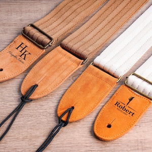 Könnte beinhalten: Nahaufnahme von drei Gitarrengurten in verschiedenen Farben. Einer ist beige mit den Initialen "HK" und einem Namen, ein weiterer ist braun und der dritte ist weiß. Jeder Gurt hat ein Lederende und eine Metallschnalle.