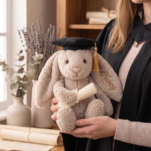 May include: A soft, beige bunny toy wearing a black mortarboard hat with a gold tassel. The bunny holds a rolled-up scroll and has the name "Nancy" embroidered on its ear. A graduation gift.