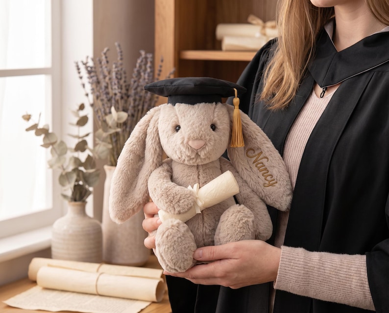 May include: A soft, beige bunny toy wearing a black mortarboard hat with a gold tassel. The bunny holds a rolled-up scroll and has the name "Nancy" embroidered on its ear. A graduation gift.
