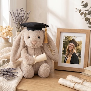 May include: A soft, light brown bunny toy wearing a black graduation cap with a gold tassel and holding a scroll. The bunny has the name "Sophie" embroidered on its ear. A framed photo of a graduate is also visible.