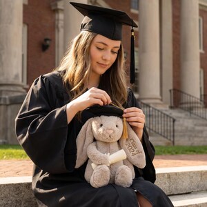 May include: A person in a black graduation gown and mortarboard adjusts the cap on a plush bunny holding a scroll. The bunny is light brown with "Class of 2024" embroidered on its ear. The background features a brick building and white columns.