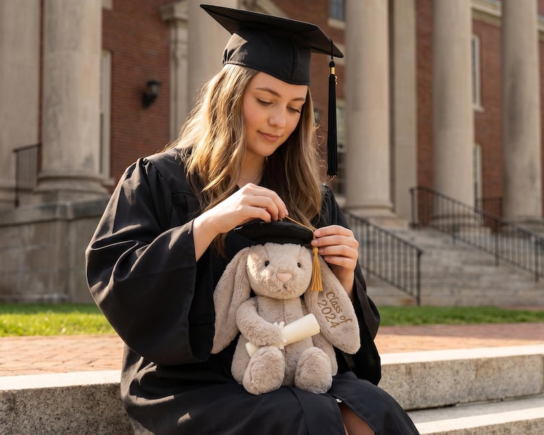 May include: A person in a black graduation gown and mortarboard adjusts the cap on a plush bunny holding a scroll. The bunny is light brown with "Class of 2024" embroidered on its ear. The background features a brick building and white columns.