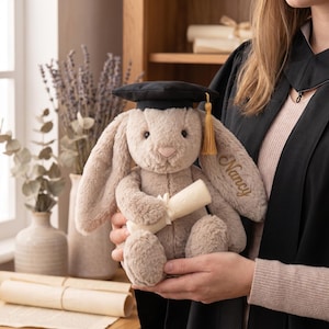 Könnte beinhalten: Ein Plüschhase mit Abschlusskappe und Schriftrolle. Der Hase ist hellbraun mit langen Ohren und mit dem Namen "Nancy" bestickt. Die Kappe ist schwarz mit einer goldenen Quaste. Ein Geschenk zur Graduierung.