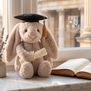 May include: A soft, beige bunny toy wearing a black mortarboard hat with a gold tassel. The bunny holds a scroll and has the name "Sophia" embroidered on its ear. A vase of lavender and an open book are nearby.