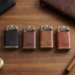 May include: Four personalized leather lighters in black, brown, and tan hues, with gold-colored metal accents. Each lighter features custom text. A cigar and a pocket watch are also visible on a wooden surface.