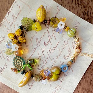 Puede incluir: Una pulsera artesanal con una variedad de cuentas coloridas en amarillo, verde, azul y marrón. La pulsera incluye cuentas con forma de flor, hoja y fruta, con una cadena y un cierre dorados. La pulsera está sobre papel vintage.