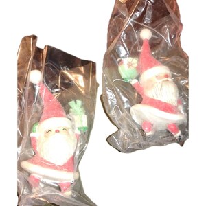 Könnte beinhalten: Zwei Weihnachtsschmuckstücke mit Weihnachtsmann-Motiv in klarer Plastikverpackung. Jedes Ornament hat einen roten Hut und Mantel, einen weißen Bart und Besatz sowie ein grünes Geschenk. Die Ornamente haben ein Vintage-Design.