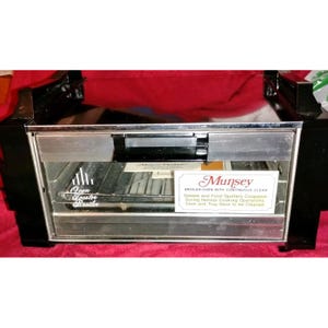 Puede incluir: Un horno-parrilla negro y plateado vintage con el texto "Munsey" y "Broiler Oven with Continuous Clean". El horno tiene una puerta de cristal y una rejilla metálica en el interior. El texto indica que la grasa y las salpicaduras de comida desaparecen durante la cocción normal.
