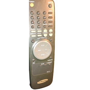 May include: A dark gray Samsung remote control with multiple buttons, including power, eject, menu, and play. The remote has a circular control pad and the Samsung logo at the bottom.