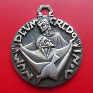 May include: A silver-toned religious pendant with the words "NUM.DEUM.CREDO.IN.V.I.A." encircling a central relief. The relief depicts two figures, one above the other, with outstretched arms. The pendant has a hexagonal loop at the top.