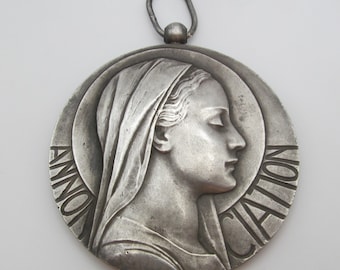 Puede incluir: Un colgante de plata con un relieve del rostro de una mujer. El colgante es redondo y tiene la palabra "ANNUNCIATION" inscrita alrededor del borde.