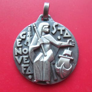 May include: A silver-toned pendant with a figure holding a staff and a ship. The pendant has the words "GENOVEFA" and "STA" in relief. The background is a solid red.