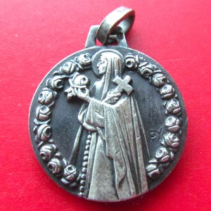 May include: A round silver-toned metal pendant featuring a relief of a figure in a hooded robe holding a cross and a flower. The figure is encircled by a wreath of small sculpted roses. A metal loop is attached to the top for hanging purposes.