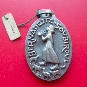 May include: Oval-shaped silver-toned pendant featuring a religious figure and the words "BERNADETTE SOUBIROU". The pendant has a small tag attached with the text "MADE IN FRANCE". The background is red.