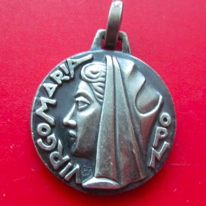 May include: A circular silver-toned metal pendant featuring a relief profile of a person wearing a head covering. The text VIRGO MARIA is embossed along the left edge, and the letters OPN are embossed on the right side. A metal loop is attached to the top.