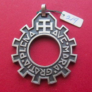 May include: A silver-toned metal pendant shaped like a gear with a central circular cutout. The text "AVE MARIA GRATIA PLENA" is embossed along the circular frame, topped with a cross symbol and a small bail. A white tag with "219" is attached.