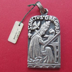 May include: A silver-toned metal pendant featuring a relief of a figure in a mitre writing at a desk, with a dove above and the text SANCTUS GREGORI. A green string attaches a small rectangular tag with the text MADE IN FRANCE to the top loop.