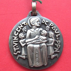 May include: A silver-toned pendant featuring a central figure holding a child, surrounded by other figures. The words "ST. VINCENT DE PAUL PPN" are inscribed around the edge. A green string is attached at the top.