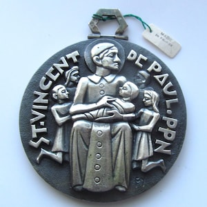 May include: A silver-toned metal medallion with a raised image of Saint Vincent de Paul holding a child. The medallion is surrounded by a black background with the words "ST. VINCENT DE PAUL. PPN" inscribed around the edge. The medallion is made in France.