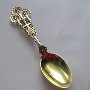 May include: A decorative metal spoon featuring a gold-toned bowl and a handle with a silver and gold finish. The handle top displays three circular motifs with flame-like details, a central cross, and a stylised geometric base design.