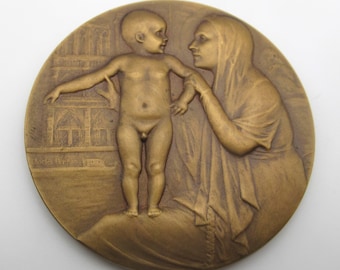 Puede incluir: Una medalla de bronce que representa a una mujer y un niño. La mujer lleva una túnica larga y sostiene la mano del niño. El niño está de pie en una plataforma y mira a la mujer. La medalla está inscrita con el nombre "Charles Perron".