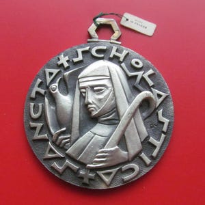 May include: A silver-toned religious medal with a depiction of a robed figure holding a staff and a dove. The medal has an ornate border with stylized lettering and a loop for hanging. The background is a solid red.