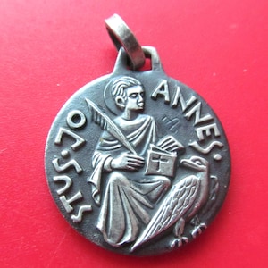 May include: A round silver-toned metal pendant featuring a relief of a figure holding a quill and a book with a cross symbol. An eagle stands to the right of the figure. The text STUS JO ANNES is embossed along the curved edge of the pendant.