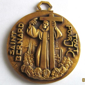 May include: A bronze-colored Saint Bernard medal. The medal features a relief of a monk holding a cross, with the words "SAINT BERNARD" and "POUR TOUT J.C." inscribed around the edge. The medal has a loop at the top for hanging.
