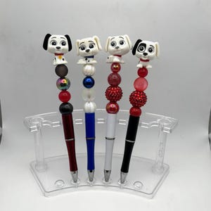 May include: Four pens with dog-themed toppers displayed in a clear acrylic stand. Each pen features a unique combination of colorful beads, including red, black, blue, and white spheres, paired with a cartoon-style puppy figure at the top of the barrel.