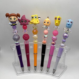 May include: A collection of six colorful beaded pens, each topped with a different character figurine. The pens feature various colors, including purple, red, orange, black, pink, and light purple, with silver tips and clear acrylic stands.