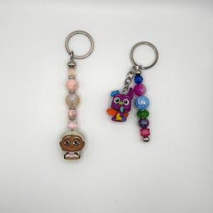 Puede incluir: Dos llaveros con cuentas y dijes coloridos. Un llavero presenta una hebra de cuentas rosa y blanca con un dije de personaje de dibujos animados. El otro llavero tiene una hebra de cuentas moradas y azules con un dije de búho de dibujos animados.