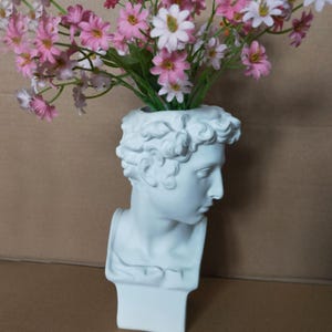 Op de afbeelding: Een witte bustevormige vaas met een klassiek ontwerp, met roze en witte bloemen. De vaas is een sculptuur van het hoofd en de schouders van een man. De bloemen voegen een vleugje kleur toe aan de neutraal getinte vaas.