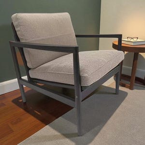 Könnte beinhalten: Ein moderner Sessel mit grauem Holzrahmen und hellbeigen Kissen. Der Stuhl hat ein rechteckiges Design mit Armlehnen und einer bequemen Sitzfläche und Rückenlehne. Der Stuhl steht auf einem hellgrauen Teppich.