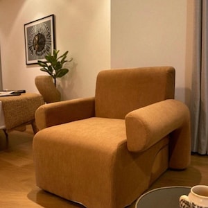 May include: A corduroy-textured, orange-brown armchair with rounded arms. The chair has a low profile and a boxy shape. The background includes a framed artwork, a small plant, and a side table. The overall aesthetic is modern and comfortable.
