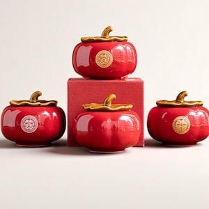 Puede incluir: Cuatro tarros de cerámica roja con tapas doradas y un emblema dorado. Los tarros están dispuestos sobre una superficie blanca, con un tarro apilado sobre una caja roja. Los tarros tienen un acabado brillante y forma de calabaza.
