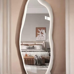 Könnte beinhalten: Ein Ganzkörperspiegel mit einem weißen, organisch geformten Rahmen. Der Spiegel spiegelt ein Schlafzimmer mit Bett, Kunstwerken und einer kleinen Ottomane wider. Der Rahmen hat eine glatte Oberfläche und verleiht der Einrichtung eine moderne Note. Höhe: ca. 170 cm.