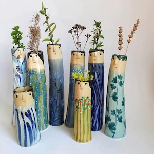 May include: A selection of handmade ceramic vases, each with a unique human face design and elongated form. The vases are painted in shades of blue, green, and yellow, with some featuring floral patterns. Each vase contains dried flowers or greenery.