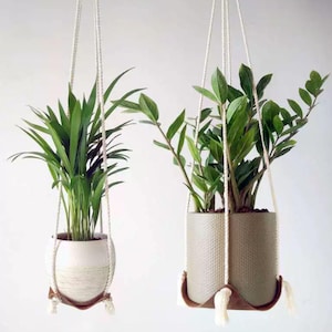 以下が含まれることがあります： 緑の植物が入った2つのハンギングプランター。左側のプランターは、織り模様と茶色の革のベースが付いた白です。右側のプランターは、織り模様と茶色の革のベースが付いた薄緑色です。どちらも白いロープで吊り下げられています。