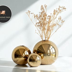 Könnte beinhalten: Drei goldene kugelförmige Vasen in verschiedenen Größen. Die größte Vase enthält getrocknete, beige farbene Blumen. Die Vasen haben eine reflektierende, metallische Oberfläche. Das Bild enthält ein Logo mit der Aufschrift "COZY HOME FINDS LIVING ROOM ACCESSORIES."