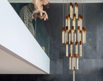 May include: A modern chandelier with multiple tiers of cylindrical, crystal-like elements. Each tier is accented with a brown, rectangular cap. The fixture hangs from thin wires, creating a cascading effect. The background features a dark gray wall.