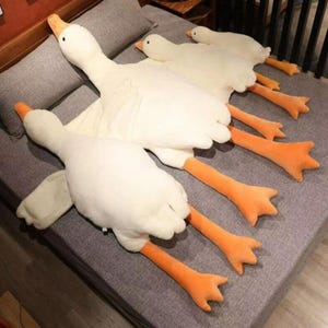 May include: A collection of plush white goose pillows with orange feet and beaks, arranged on a grey surface. The pillows are soft and fluffy, with outstretched necks and wings. The geese are lying down.