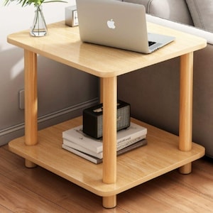 May include: A light wood square side table with a lower shelf. The table has four rounded legs and a natural wood grain finish. A laptop, books, and a small speaker are on the table. A vase with a plant is on the top.