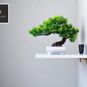 May include: A small, artificial bonsai tree with vibrant green foliage and a dark brown trunk sits in a white rectangular pot. The tree is on a white shelf, next to a small black decorative cup. The shelf is supported by gold-colored brackets.