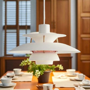 Puede incluir: Lámpara colgante blanca con un diseño en capas en forma de platillo. La luz cuelga sobre una mesa de comedor con tazas y platillos blancos. El accesorio tiene una estética limpia y moderna.