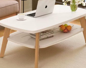 May include: A white rectangular coffee table with a lower shelf and wooden legs. A laptop, a cup and saucer, a bowl of fruit, and magazines are on the table. The table is on a light-colored rug.