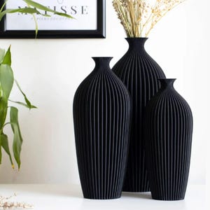 Puede incluir: Tres jarrones decorativos negros con un diseño acanalado. Los jarrones son de diferentes tamaños y se apoyan sobre una superficie blanca. Un jarrón contiene flores secas. Una impresión enmarcada es visible en el fondo.