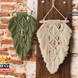 May include: Two macrame wall hangings with wooden dowels and wooden beads. The wall hanging on the left is green and the wall hanging on the right is cream.